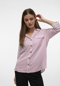 Camisa de botones rayada en rosa y blanco, con dos bolsillos frontales, cuello y mangas largas remangadas en los puños. Textura de tela suave.