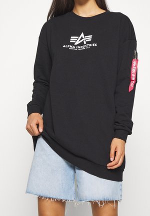 Osoba ubrana w czarną long-sleeve bluzę Alpha Industries z białym logo i czerwoną metką na rękawie, w połączeniu z jasnoniebieskimi szortami dżinsowymi.