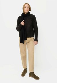 Ciemny sweter z dzianiny z wysokim kołnierzem, w połączeniu z beżowymi spodniami i zielonymi botkami za kostkę. Elegancka czarna chusta jest zarzucona na jedno ramię.