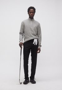 Άνδρας που φορά γκρι πουλόβερ και μαύρο παντελόνι κρατά μπαστούνι του γκολφ με λευκό γάντι κρυμμένο στη μέση σε λευκό φόντο.