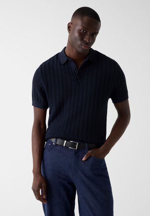 Młody mężczyzna ubrany w granatową, krótko rękawową dzianinową koszulkę polo, ciemne jeansy i czarny pasek, z jedną ręką w kieszeni, na jednolitym tle.