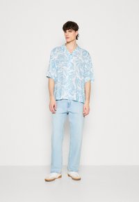 Camicia azzurra a maniche corte con un motivo ondulato bianco, abbinata a jeans azzurri e sneakers bianche con suola beige.