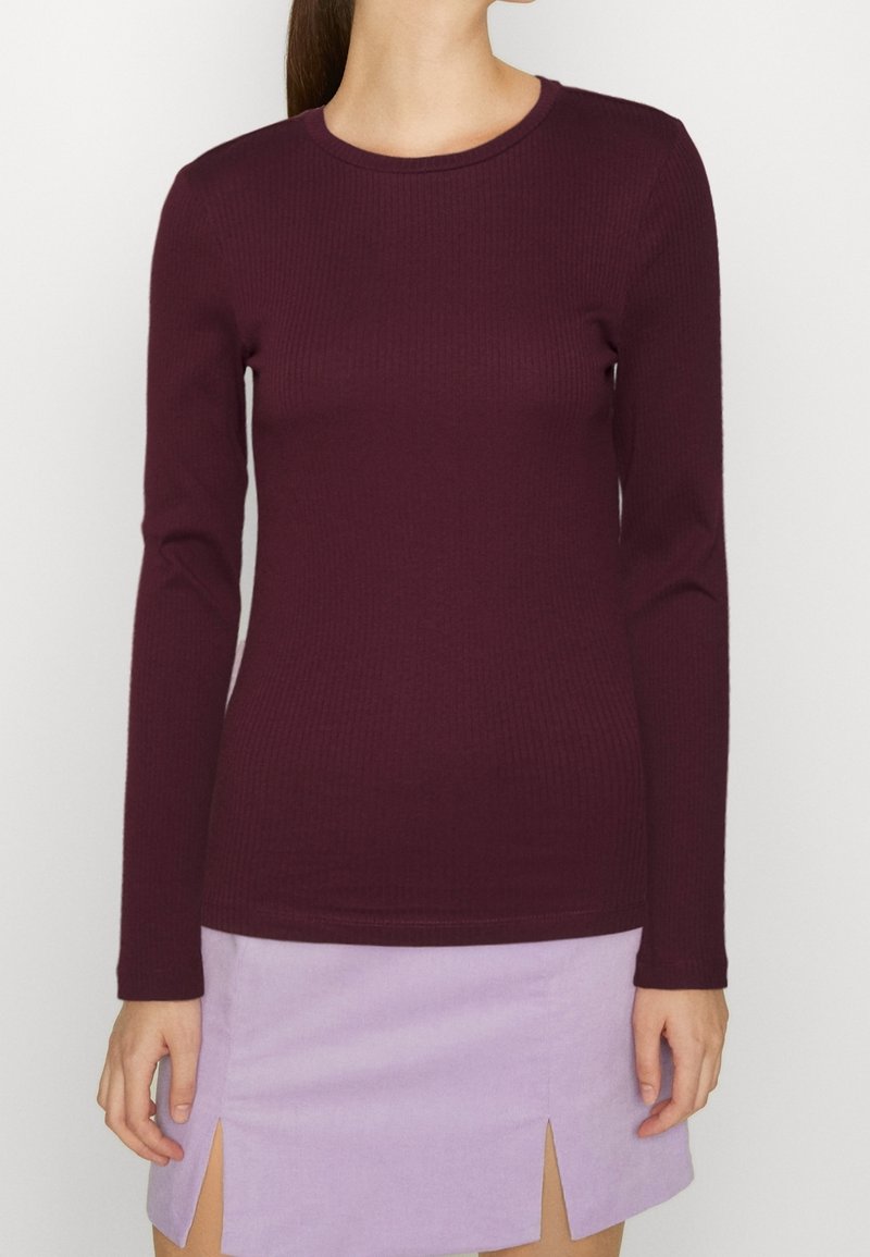 Femme portant un haut à manches longues ajusté en ribbed de couleur violet foncé et une mini-jupe violet clair avec des fentes à l'avant, se tenant devant un fond uni.