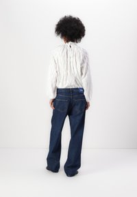 Hvit teksturert bluse med samlet bak og denim bukser med vide ben som har en blå etikett og to baklommer. Enkel bakgrunn.