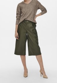 Pull en tricot marron avec col en V et texture côtelée, associé à un pantalon court vert olive avec fermeture à bouton sur le devant et jambes larges.