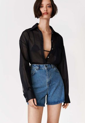 Kobieta ubrana w przezroczystą czarną koszulę na guziki, na czarnym biustonoszu typu braletka, w połączeniu z wysokim stanem niebieskich szortów dżinsowych na jednolitym tle.