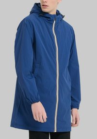 Veste en nylon bleu avec une capuche à cordon, des poignets élastiques, des poches avant et une fermeture éclair intégrale présentant une bande rayée au centre.