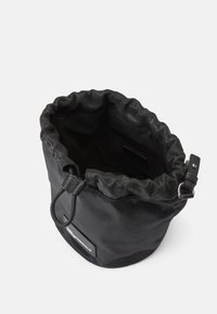 Borsa a secchiello nera in tessuto con apertura a coulisse, superficie texture, tracolla regolabile e etichetta del marchio visibile sul lato. L'interno mostra profondità.