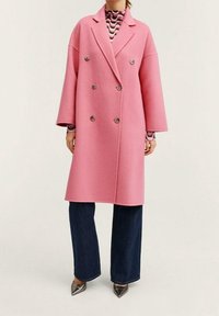 Dvojřadový růžový kabát z vlny, s širokými rukávy, šesti knoflíky a rovný střih, v kombinaci s tmavými rozšířenými džíny a špičatými botami.