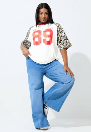 Kobieta pozująca, ubrana w białą koszulę z czerwonym numerem „89” i rękawami w panterkę, szerokie niebieskie dżinsy oraz czarno-białe trampki.