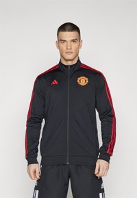 Sort sportsjakke med røde striber og Manchester United-logo. Har lynlås foran og højt krave. Bæres med matchende sorte bukser.