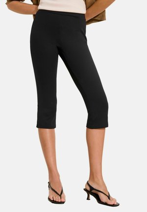 Frau trägt schwarze knappe, schmale Hosen kombiniert mit schwarzen Sandalen mit Absatz, steht mit den Händen hinter dem Rücken.