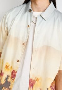 Košile s krátkým rukávem ve světlé béžové barvě, s potiskem koně ve stylu akvarelu na spodní straně. Zapínání na knoflíky s límečkem, vyrobená z měkké látky.
