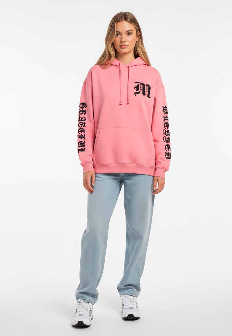 Felpa rosa con tasca frontale, cappuccio con lacci e scritte nere sulle maniche. Abbinata a jeans azzurri chiari e sneakers bianche.
