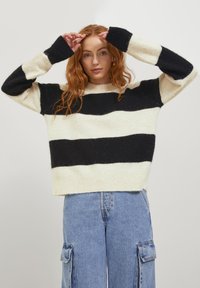 Gestreifter Strickpullover in Creme und Schwarz, mit langen Ärmeln und gerippten Bündchen. Das Material wirkt weich mit einer gemütlichen Textur und hat eine figurbetonte Passform.