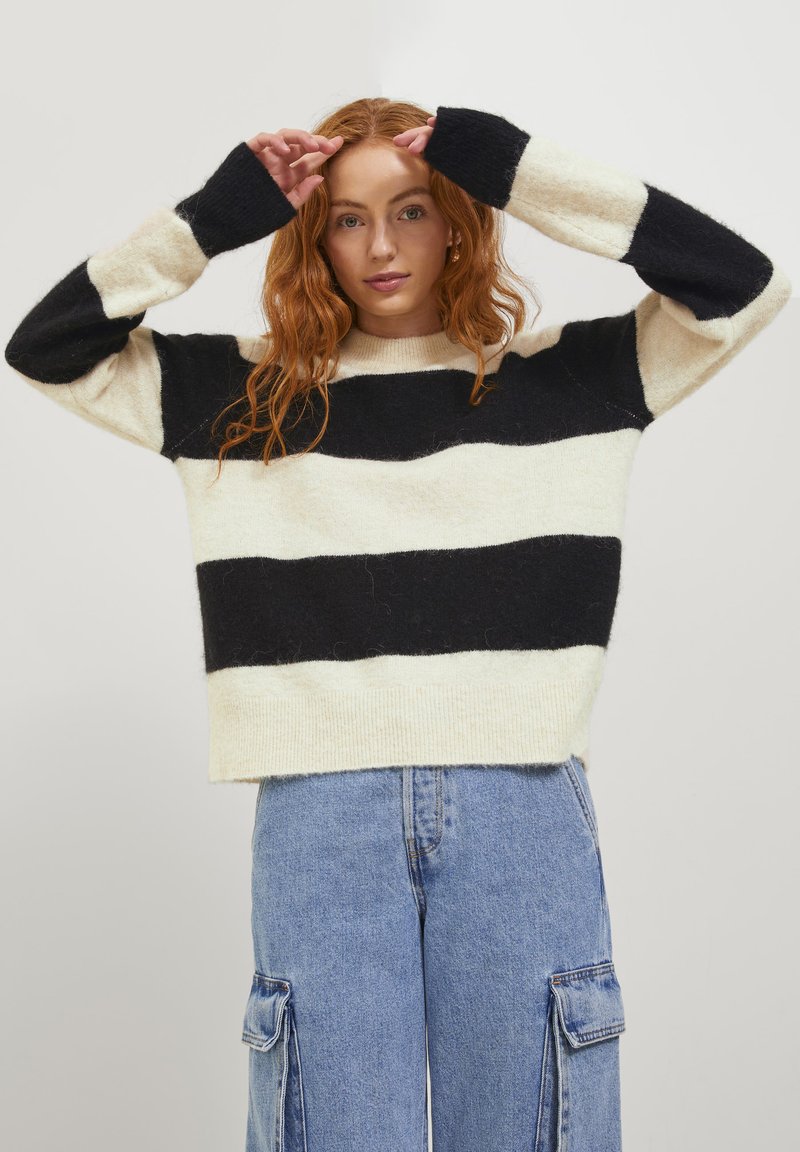 Gestreifter Strickpullover in Creme und Schwarz, mit langen Ärmeln und gerippten Bündchen. Das Material wirkt weich mit einer gemütlichen Textur und hat eine figurbetonte Passform.