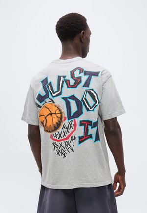 Hátrafelé néző férfi szürke pólóban, amelyen színes "JUST DO IT" felirat és egy kosárlabda grafika látható, amely áthalad a háton lévő gyűrűn.