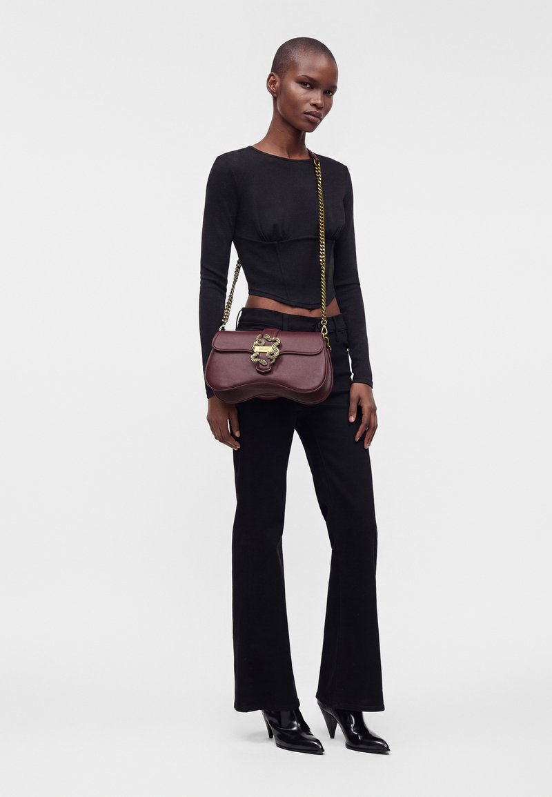 Femme portant une tenue entièrement noire avec des bottines à talons aiguilles pointues, portant un sac à épaule marron avec une fermeture dorée décorative et une bandoulière chaîne.