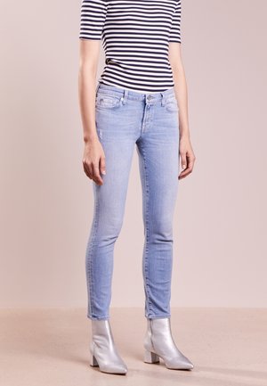 Persoon draagt lichtblauwe skinny jeans, een zwart-wit gestreept T-shirt met korte mouwen en zilveren enkellaarsjes, staand op een beige vloer.