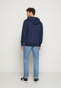 Marineblauer Kapuzenpullover mit geripptem Saum, getragen mit hellblauen Jeans und schwarzen Schuhen. Die Ansicht zeigt Rücken- und Seitenprofil.