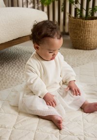 Kind trägt ein cremefarbenes Langarmshirt und einen weißen Tüllrock, sitzt auf einer strukturierten, hellen Matte mit sichtbaren nackten Füßen.