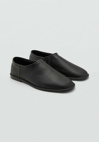Zapatos de cuero negro sin cordones con un acabado texturizado, punta redondeada y suela de perfil bajo. Diseño minimalista sin hardware visible.