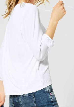 Camicia bianca a maniche lunghe con polsini arricciati, caratterizzata da una vestibilità ampia e un tessuto texturizzato. Abbinata a jeans in denim blu.