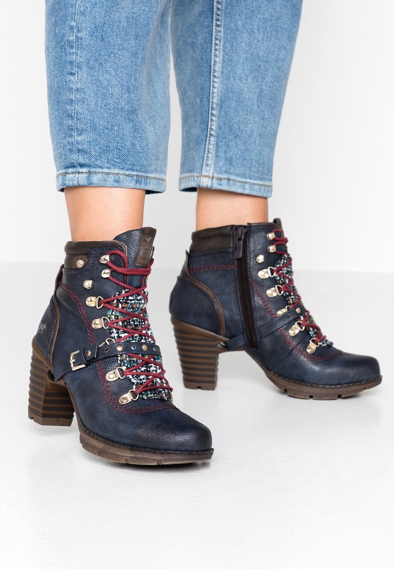 Botas de tobillo de tacón en azul marino con cordones rojos, cremallera lateral y lengüeta con estampado, combinadas con jeans cortos de color azul claro.