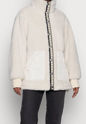 Veste polaire blanche avec col montant, fermeture à boutons-pression à l'avant, deux poches zippées frontales, portée sur des manches crème et un pantalon foncé.