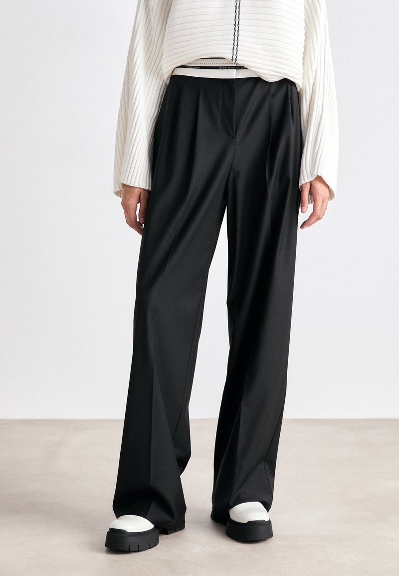 Personne portant un pantalon noir à jambes larges, un pull blanc côtelé et des chaussures épaisses noires et blanches, debout sur un sol clair devant un fond blanc.