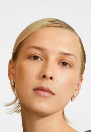 Close-up van een vrouw met teruggekamd blond haar, die gouden en diamanten creolen draagt, met een neutrale gezichtsuitdrukking.
