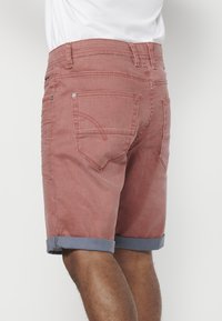 Shorts en denim rouge avec des revers bleus roulés, plusieurs poches à l'arrière et des accents de couture subtils. Design simple, coupe décontractée.