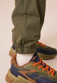 Zelené jogger nohavice so zúženými manžetami a textúrovaným materiálom, doplnené o viacfarebné tenisky s olivovými, námorníckymi a oranžovými akcentami.