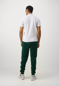 Biela polo tričko s krátkymi rukávmi, skombinované so zelenými zamotanými teplákmi s čiernymi bočnými pruhmi. Model má na sebe biele športové topánky.