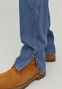 Vaqueros de mezclilla azul con un ajuste relajado, con aberturas laterales en los tobillos, combinados con botas de ante marrón.