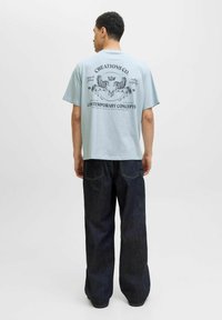 Lyseblå T-shirt med korte ærmer og grafisk print med løver og tekst på ryggen, parret med mørkeblå vide jeans.