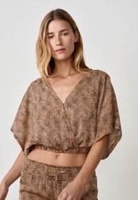 Braune, geteilte Bluse mit V-Ausschnitt, kurzen Ärmeln und einem schwarzen Spritzmuster. Leichtes Material mit lockerem Sitz. Kombiniert mit passenden Unterteilen.