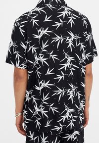 Camisa de manga corta negra con estampado de hojas de bambú blancas. Presenta un corte relajado y un cuello clásico. Tejido suave con una textura lisa.