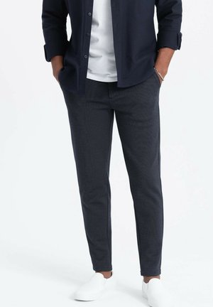 Mann trägt dunkle, strukturierte Slim-Fit-Hose, ein weißes Hemd, ein dunkles, offen getragenes Hemd, weiße Slip-On-Schuhe und ein silbernes Armband.