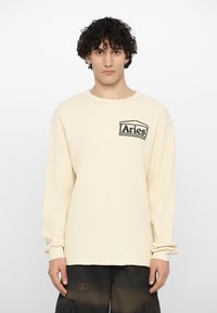 Haut à manches longues crème en tissu tricot texturé, encolure ronde et logo "Aries" noir sur le côté supérieur gauche. L'ourlet est légèrement brut.