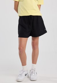 Mustat shortsit, joissa on kiristettävä vyötärö, kevyttä materiaalia ja pieni logo-yksityiskohta. Yhdistetty vaaleankeltaiseen T-paitaan ja valkoisiin lenkkareihin.