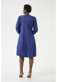 Zila, ceļgala garuma kleita ar uzpūstām piedurknēm un taisnu siluetu. Izgatavota no gludas auduma; tai ir aizmugures šuves un kabatas.