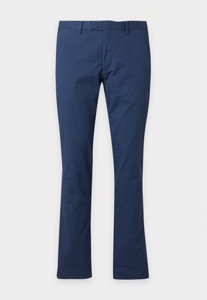 Homme portant un t-shirt vert foncé à manches courtes, un pantalon bleu marine et des baskets blanches, debout devant un fond blanc uni.