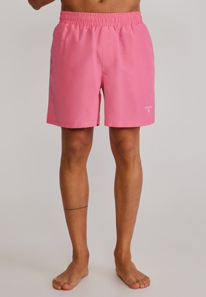 Uomo con pantaloncini elasticizzati rosa brillante, in piedi scalzo su uno sfondo chiaro e uniforme, visibili tronco inferiore e gambe.