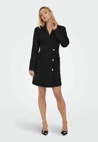 Abito blazer nero su misura con maniche lunghe, revers a punta, sei bottoni silver e due tasche frontali. Texture elegante e silhouette aderente.