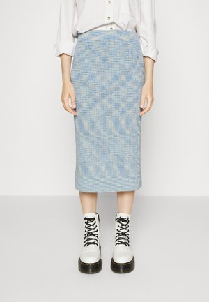 Mujer con falda de punto azul hasta la rodilla, camisa blanca abotonada con mangas remangadas y botas blancas con cordones y suelas negras.