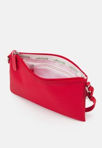 Custodia con zip rossa realizzata in materiale testurizzato. L'interno presenta una fodera bianca con il marchio verde "LACOSTE". Include una tracolla.