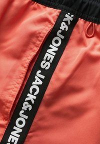 Koraalkleurige shorts met een zwart elastische tailleband. Voorzien van een zijzak en een zwart contrasterende bies met witte tekst: "JACK & JONES." Gladde textuur.