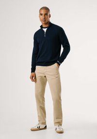 Maglione con zip colla navale in materiale morbido, pantaloni chinos beige e sneakers bianche con accenti scuri. Design semplice e pulito, senza motivi o decorazioni.