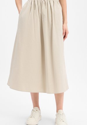 Beiger Midirock mit elastischem Bund, kombiniert mit weißen Schnürsneakern, Modell steht mit teilweise in den Taschen steckenden Händen.